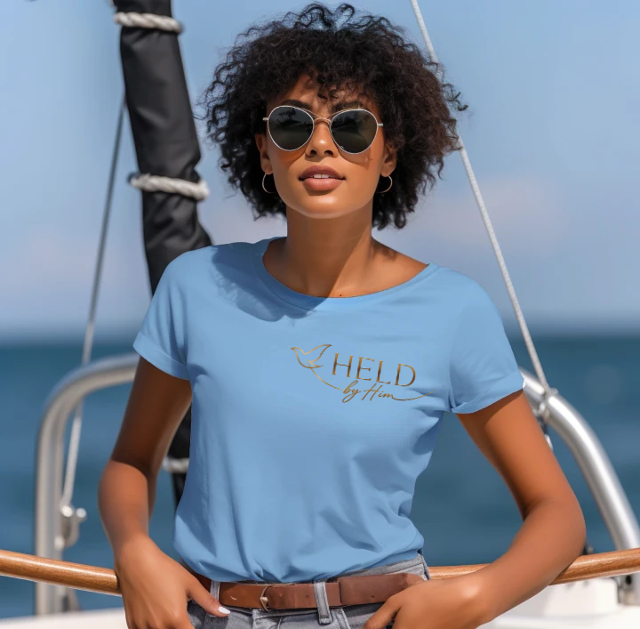 A woman with curly hair and sunglasses wearing a light blue t-shirt with a gold 'I AM HELD by Him' graphic, posing on a boat with the ocean in the background.