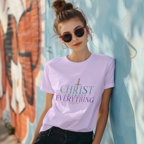 A woman wearing a light purple graphic t-shirt with the text 'CHRIST CHANGES EVERYTHING' and a sword graphic, posing against a blue graffiti wall.